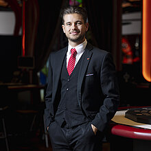  Key Account Manager im Casino Graz, Aris Konstantoupulos, trägt einen schwarzen Anzug mit roter Krawatte und steht an einem Pokertisch, lächelnd in die Kamera.